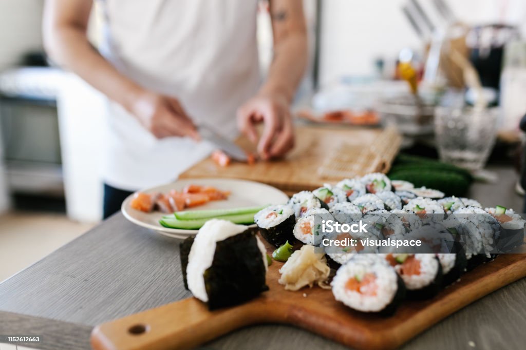 Une femme mélangeant des ingrédients et préparant des sushis sur un plan de travail en cuisine, avec des ingrédients comme du riz, du saumon et des légumes étalés autour d'elle."