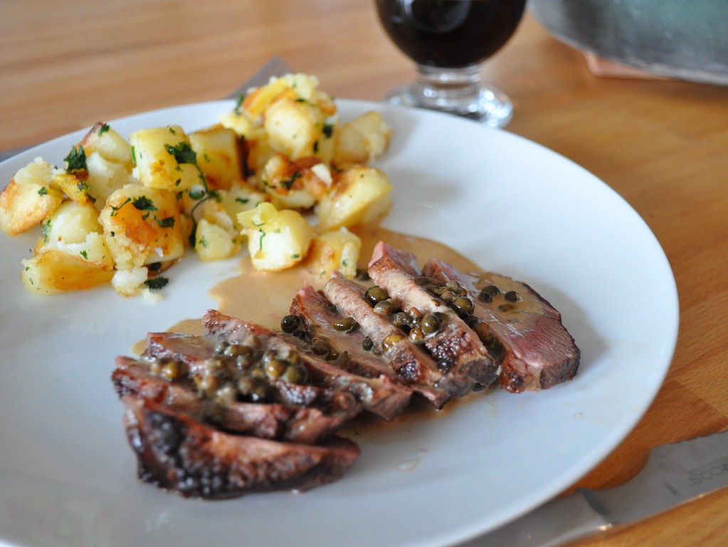 Assiette de magret de canard tranché, nappé d'une sauce aux câpres, accompagné de pommes de terre sautées et d'un verre de boisson brune."