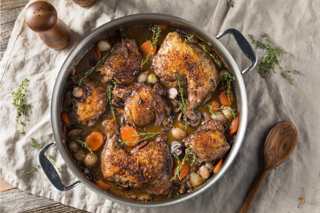 alt="Coq au vin traditionnel servi dans une cocotte en fonte avec carottes, champignons et sauce au vin rouge"